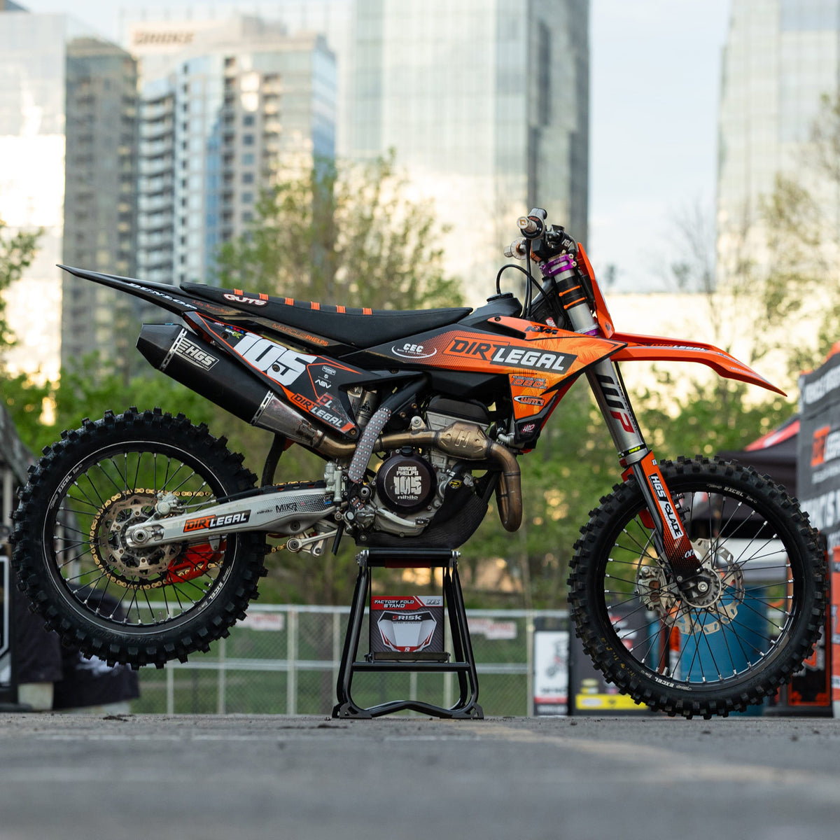 Moto tout-terrain orange et noire sur un Risk Racing F.F.S. se tenir debout avec un paysage urbain en arrière-plan