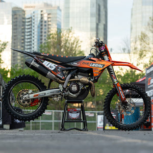 Moto tout-terrain orange et noire sur un Risk Racing F.F.S. se tenir debout avec un paysage urbain en arrière-plan